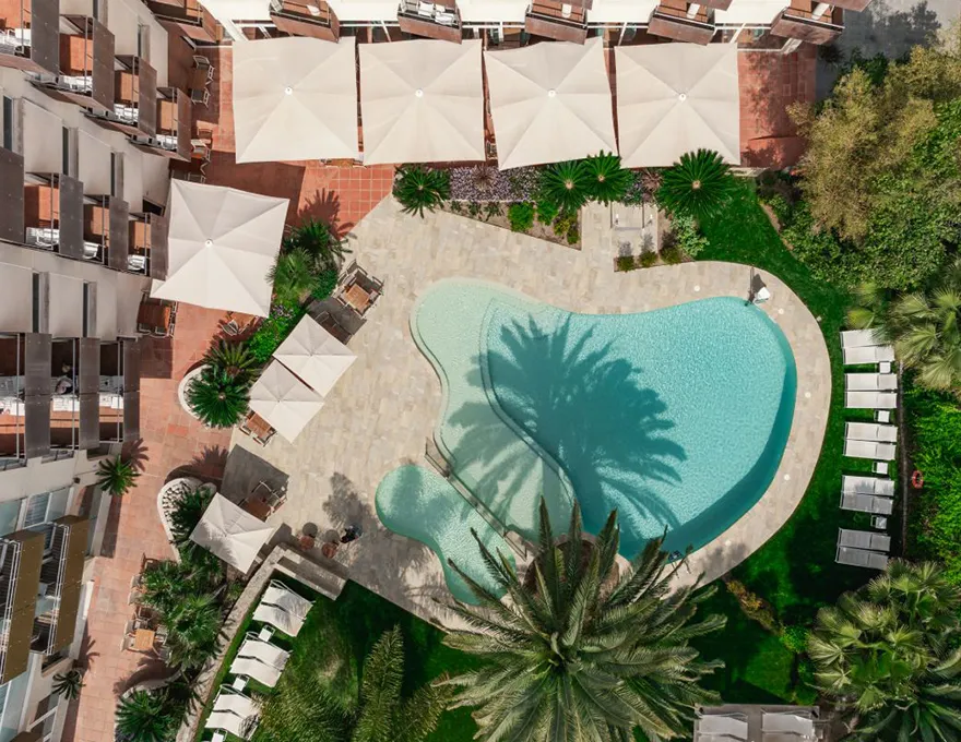 Piscina de forma irregular rodeada de sombrillas y tumbonas vista desde arriba.