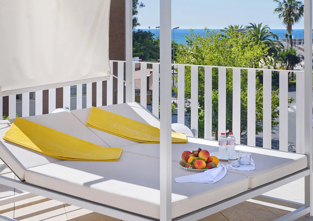 Cama con toallas amarillas, frutas y agua, vista al mar en terraza soleada.