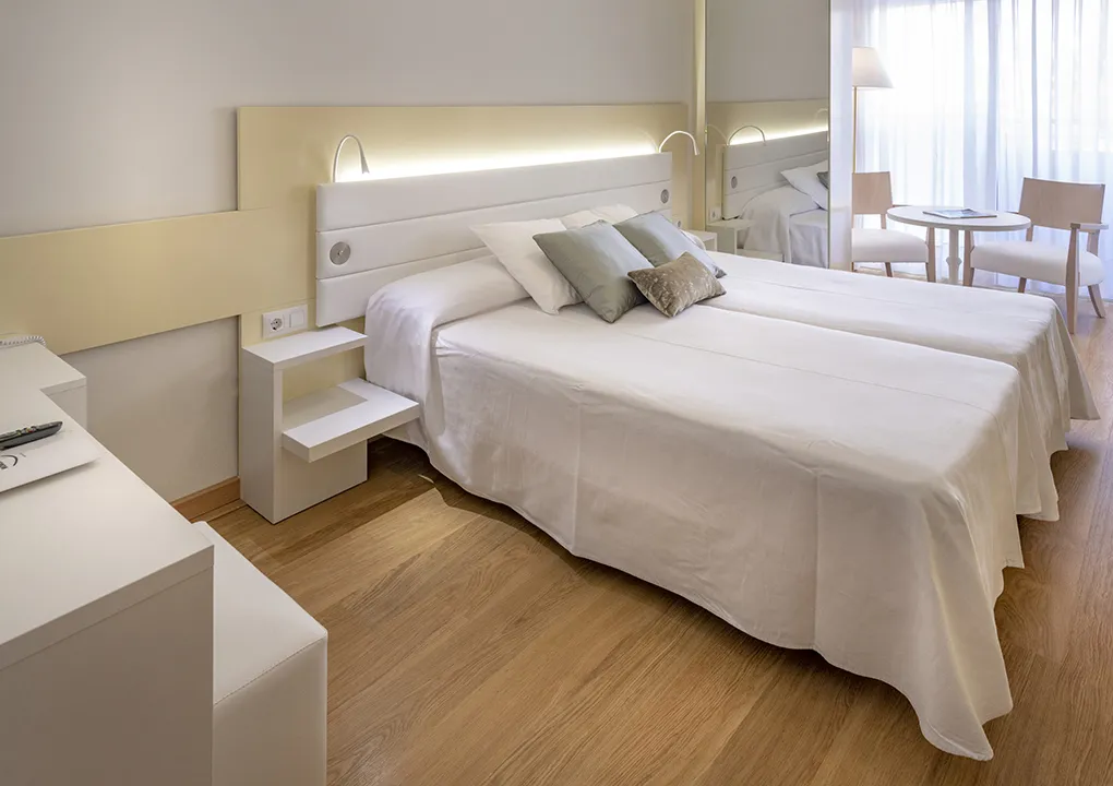 Habitaci&oacute;n de hotel con dos camas, mesa con sillas y luz natural.