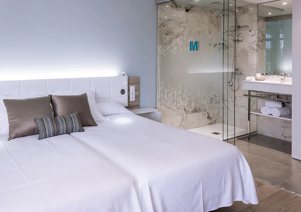 Habitaci&oacute;n de hotel moderna con cama, almohadas y ba&ntilde;o con ducha de cristal.