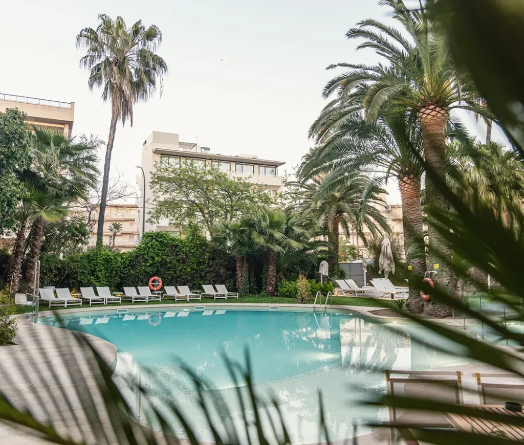 Piscina rodeada de palmeras y tumbonas vac&iacute;as en un entorno urbano.