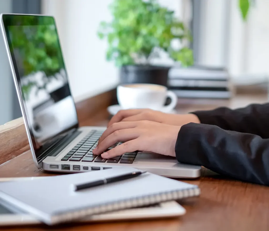 Manos escribiendo en laptop sobre mesa con cuaderno, pluma y taza al fondo.