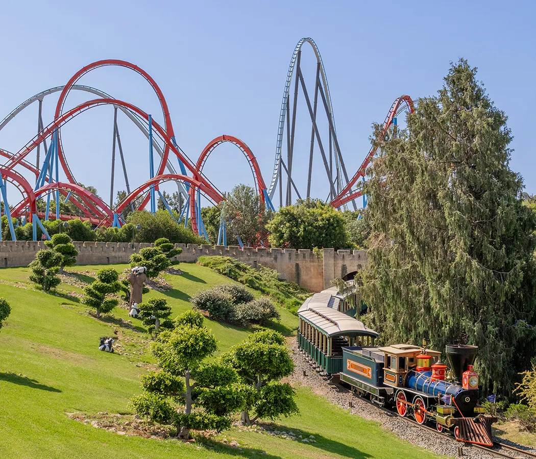 Tren en parque con monta&ntilde;a rusa al fondo y &aacute;reas verdes.