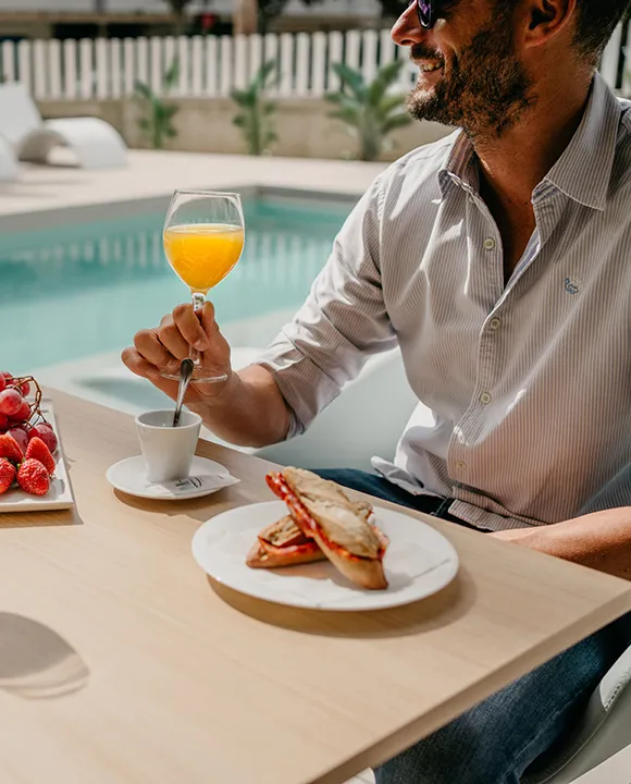 Hombre desayunando junto a una piscina con brindis y jugo naranja.
