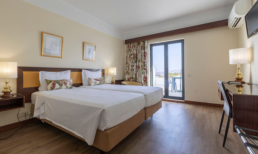 Quarto de hotel iluminado com duas camas, varanda e decora&ccedil;&atilde;o aconchegante.