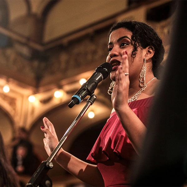 Mulher em vestido vermelho canta no microfone em ambiente iluminado.