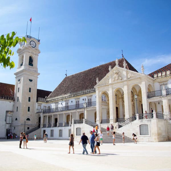 Pr&eacute;dio hist&oacute;rico com torre do rel&oacute;gio sob c&eacute;u azul, pessoas passeiam na pra&ccedil;a.