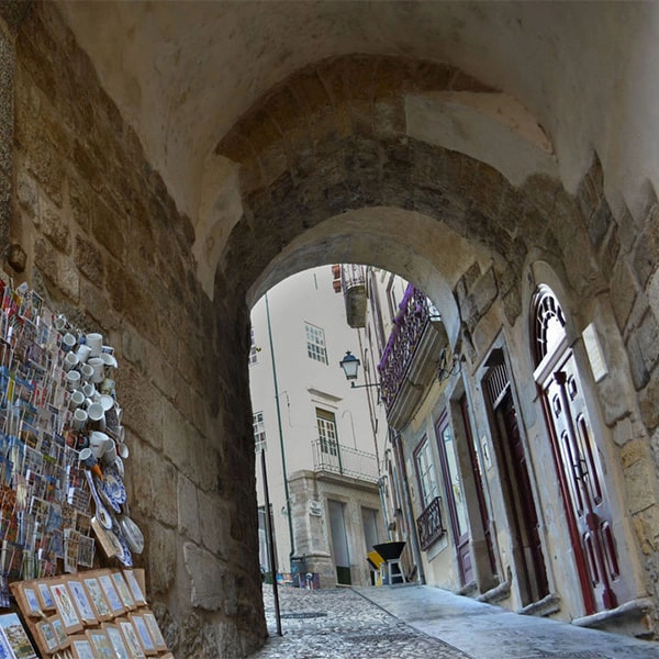 Arco de pedra em rua estreita com lojinhas e portas antigas.