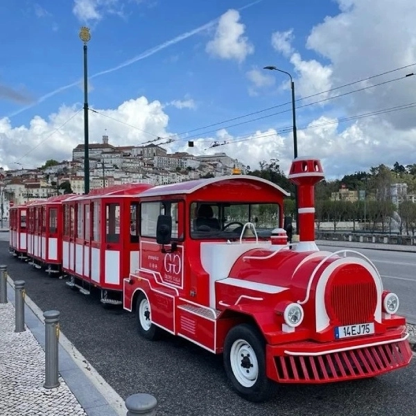 Trem tur&iacute;stico vermelho na rua com cidade ao fundo e c&eacute;u azul.
