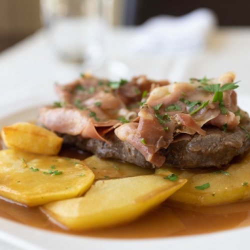 Carne com presunto e batatas, servidos com molho e ervas em um prato branco.