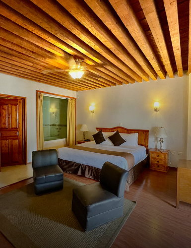 Habitaci&oacute;n con cama doble, sillas negras, techo de vigas y luz c&aacute;lida.