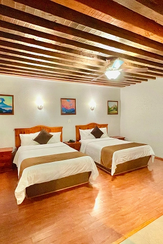 Habitaci&oacute;n con dos camas, l&aacute;mparas de pared y techo de vigas de madera.