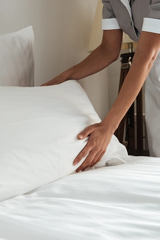 Persona arreglando una cama con s&aacute;banas y almohadas blancas.