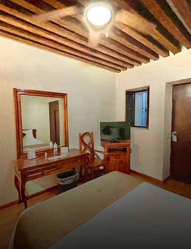 Habitaci&oacute;n con cama, escritorio, espejo y televisi&oacute;n. Techo con vigas de madera.
