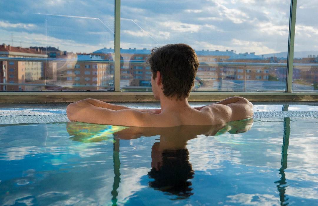 Hombre en piscina mirando por una ventana con vista urbana y cielo nublado.