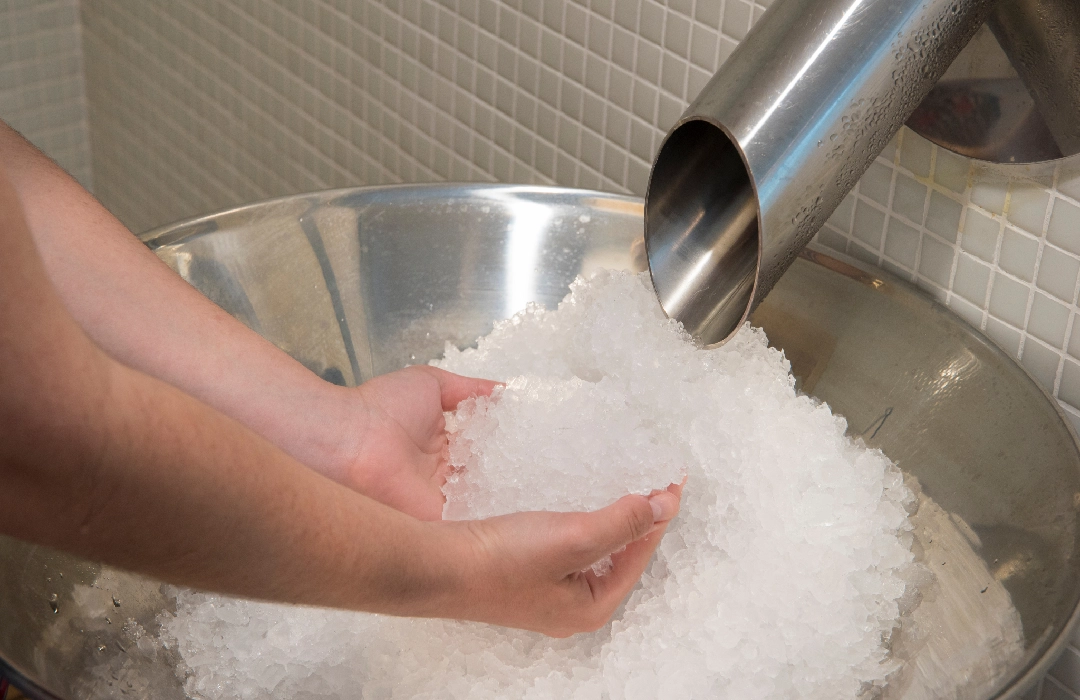 Manos recogiendo hielo en un recipiente de metal bajo un tubo dispensador.