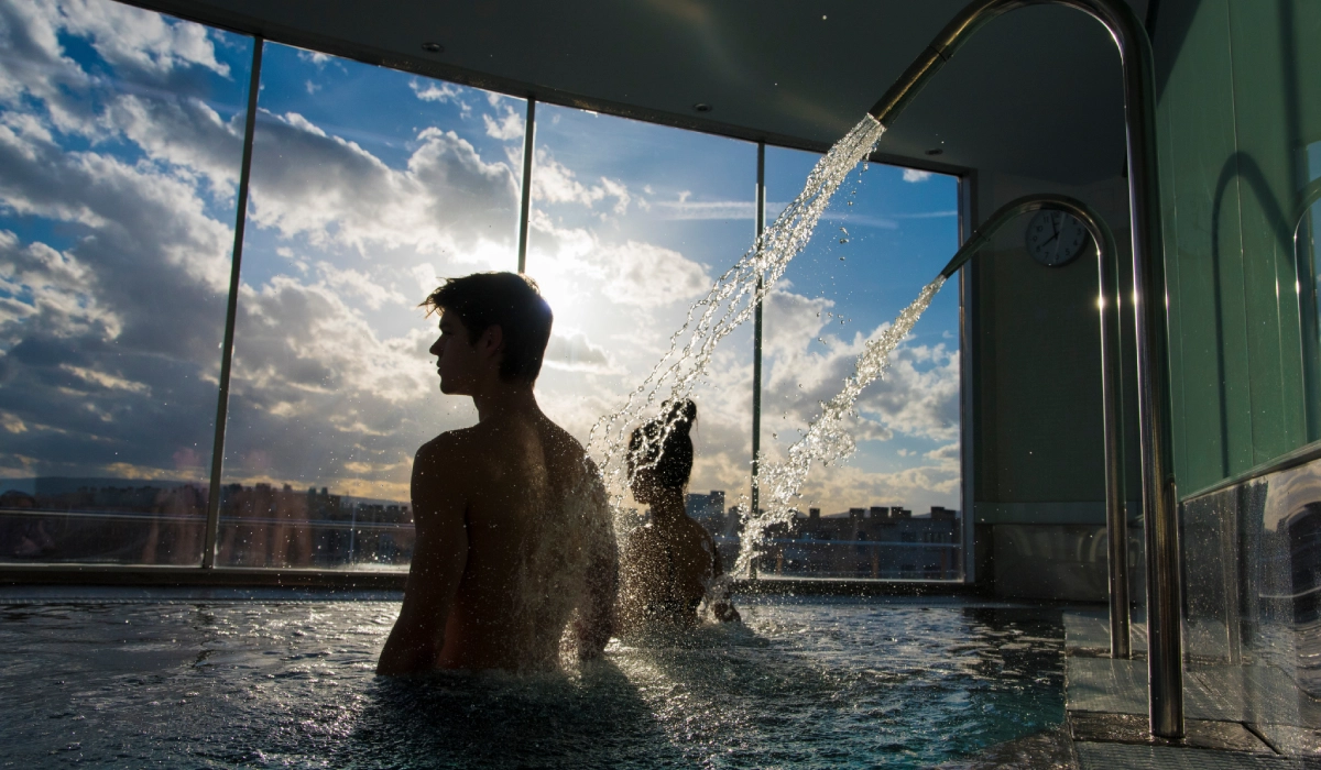 Personas en jacuzzi bajo chorro de agua, cielo nublado al fondo.