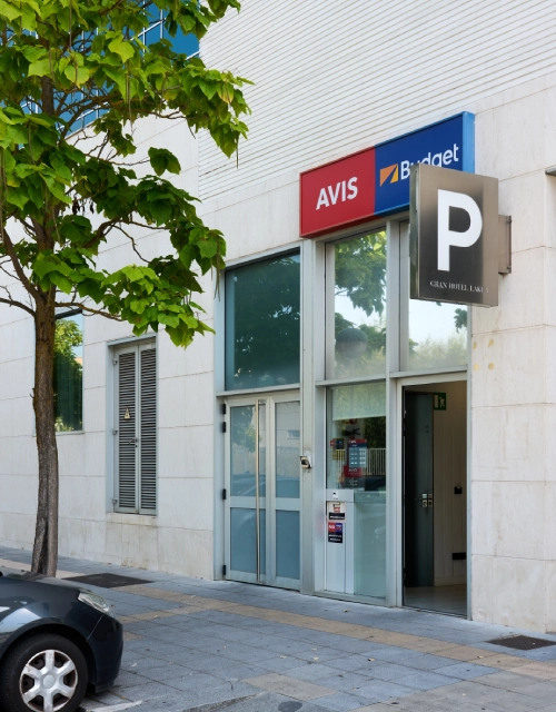 Entrada de locales de AVIS y Budget con &aacute;rbol y coche estacionado afuera.