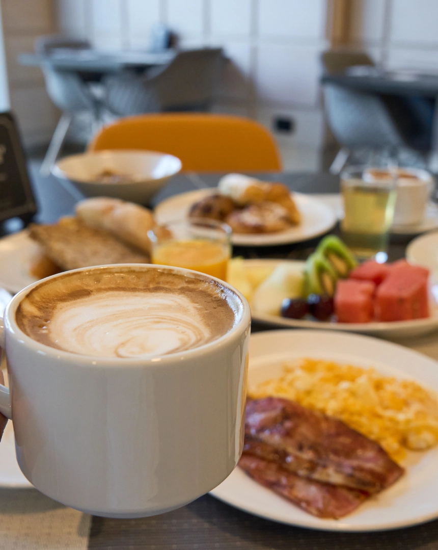 Taza de caf&eacute; con desayuno de frutas, jugo, bacon y huevos en el fondo.