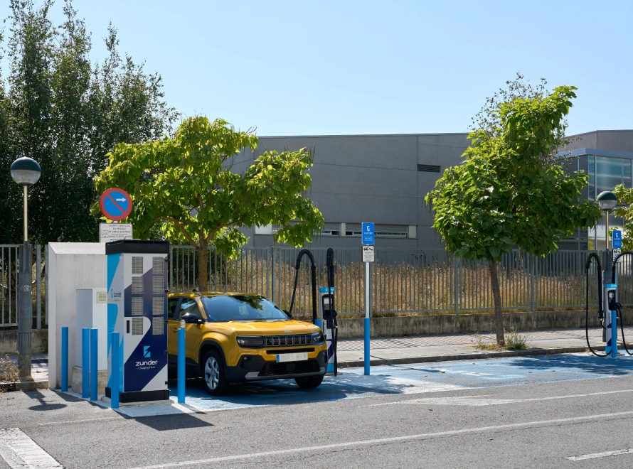 Coche amarillo cargando en estaci&oacute;n el&eacute;ctrica con &aacute;rboles alrededor.
