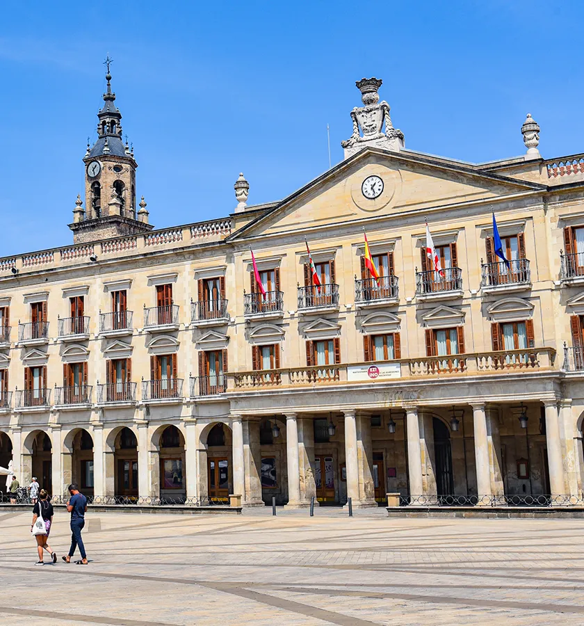 Edificio hist&oacute;rico con reloj y banderas, personas caminando frente a &eacute;l.