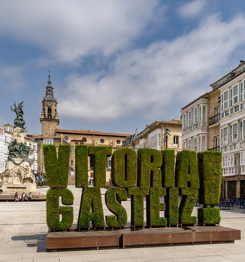Letras verdes de "Vitoria-Gasteiz" en una plaza con edificios y estatua detr&aacute;s.