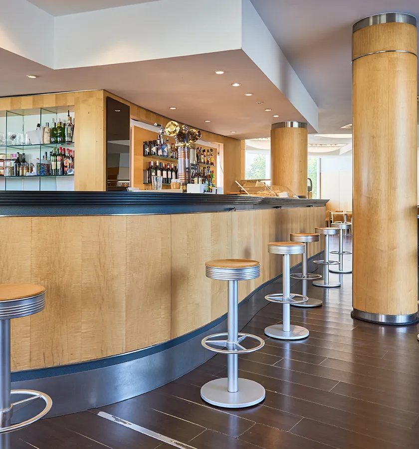 Barra de bar moderna con taburetes met&aacute;licos y estantes de botellas al fondo.