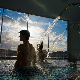 Personas en una piscina bajo un chorro de agua con vista al atardecer.