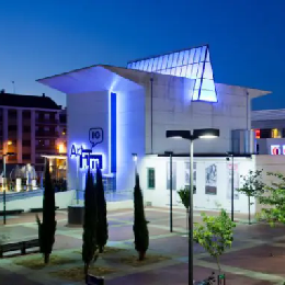 Edificio blanco iluminado de noche con &aacute;rboles y una plaza alrededor.
