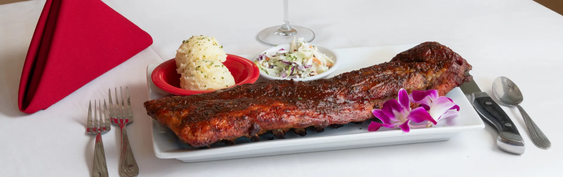 Costillar asado con puré, ensalada y flor decorativa en plato, junto a cubiertos y servilleta roja.
