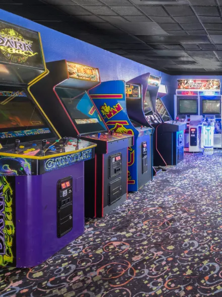 Sala de arcade retro con m&aacute;quinas de videojuegos cl&aacute;sicas alineadas sobre una alfombra colorida.