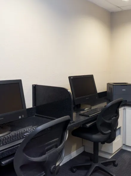 Sala de computadoras con monitores apagados y sillas negras frente a un escritorio compartido.