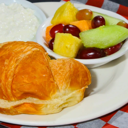 Croissant con ensalada de frutas y plato de arroz, sobre mantel de cuadros rojos y blancos.