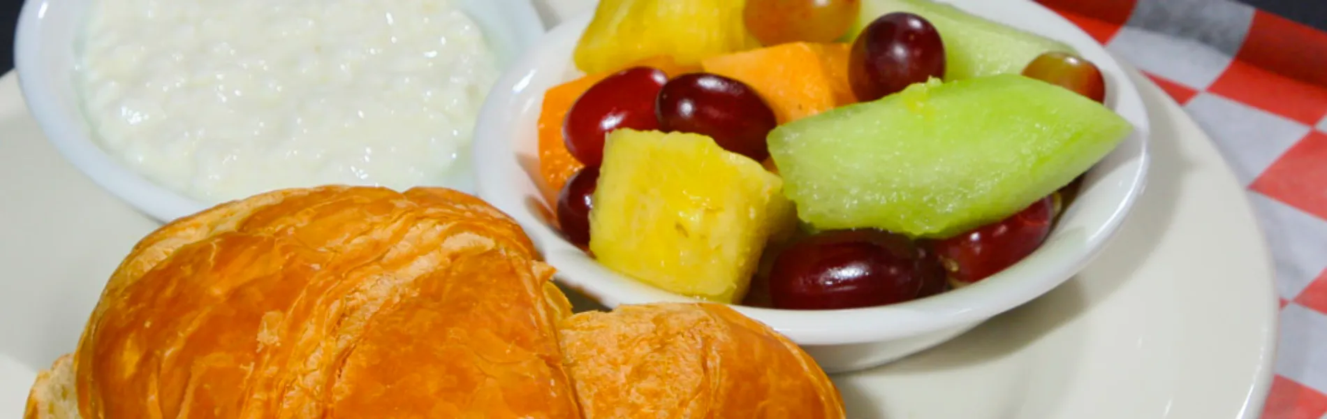 Croissant con ensalada de frutas y tazón de requesón sobre un plato blanco a cuadros.