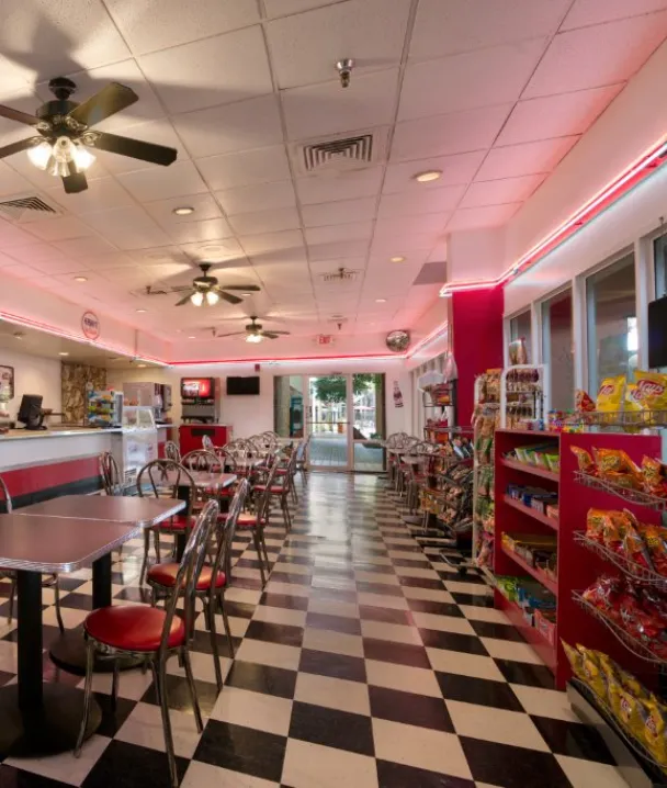 Interior de una cafeter&iacute;a estilo retro con suelos ajedrezados y sillas rojas, estantes de snacks.