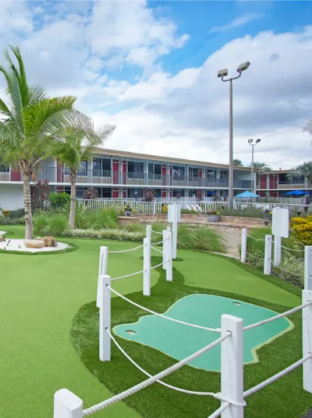 Minigolf en jard&iacute;n con palmeras, cerca de un edificio moderno bajo un cielo parcialmente nublado.