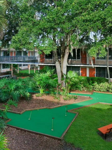 Minigolf entre &aacute;rboles y arbustos, rodeado por un edificio de dos pisos con puertas naranjas.