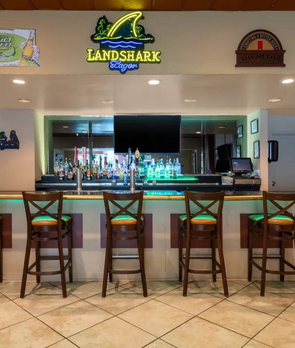 Bar con ne&oacute;n de "Landshark Lager", cuatro sillas altas de madera frente a un mostrador iluminado.