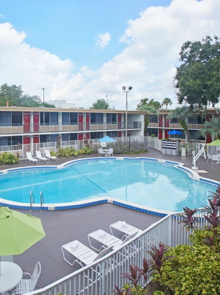Piscina exterior rodeada de tumbonas y un edificio de dos pisos en un d&iacute;a soleado.