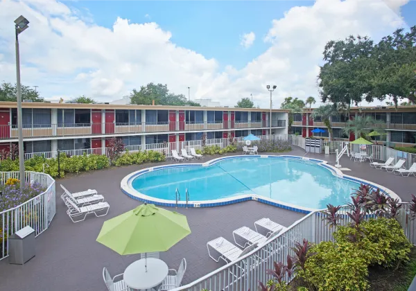 Piscina al aire libre rodeada de tumbonas, sombrillas y un edificio de dos pisos al fondo.
