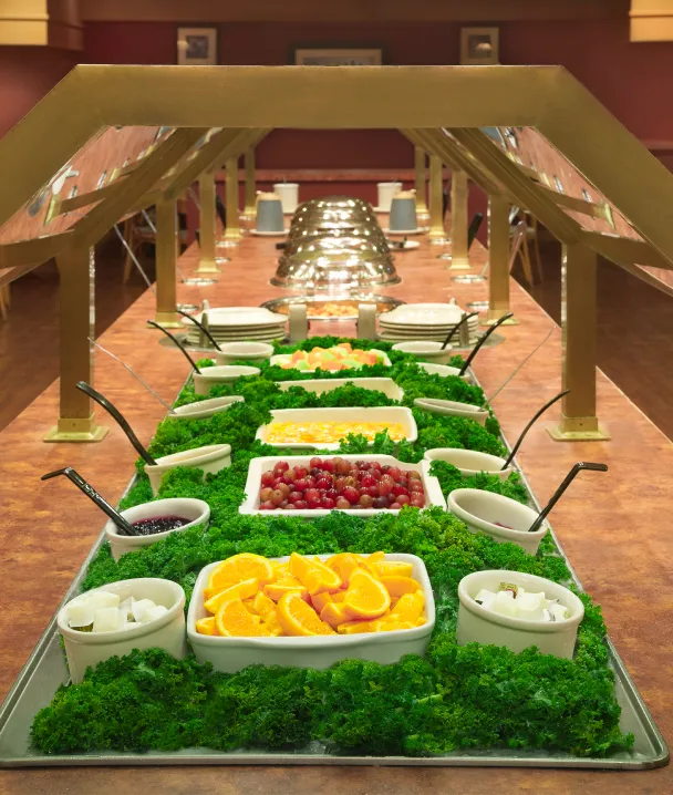 Buffet de frutas variadas en recipientes sobre una mesa, rodeados de hojas verdes decorativas.