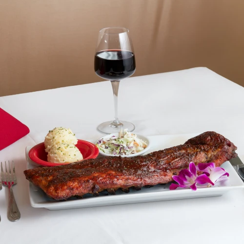 Costillar asado con puré, ensalada, orquídea y copa de vino tinto, sobre mesa de mantel blanco.