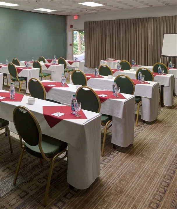 Sala de conferencias con mesas alineadas, sillas verdes, y botellas de agua sobre mantel blanco.