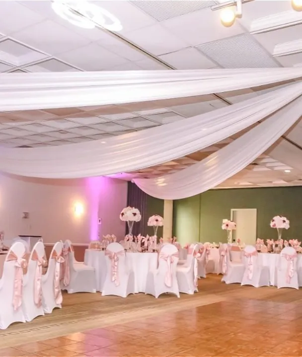 Salón decorado para evento, con mesas vestidas de blanco y rosa, y techo con telas drapeadas.