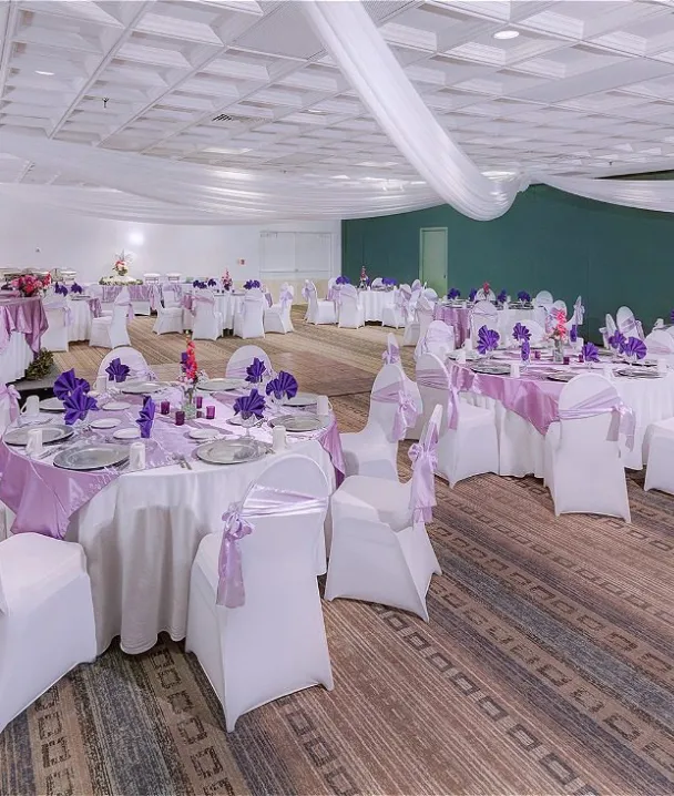 Salón de eventos decorado con manteles y servilletas púrpuras, mesas y sillas blancas.