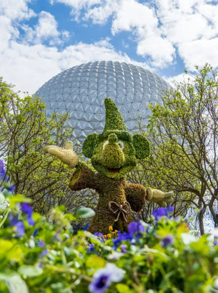 Figura de arbusto con forma de personaje, fondo de c&uacute;pula plateada y cielo despejado con flores.