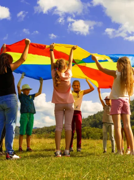 Ni&ntilde;os jugando con un paraca&iacute;das de colores sobre un campo bajo un cielo azul.