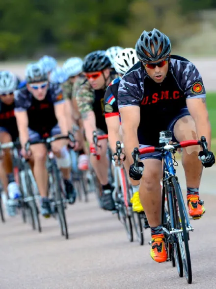 Ciclistas en fila avanzan en una carretera, liderados por un hombre con uniforme de ciclismo USMC.