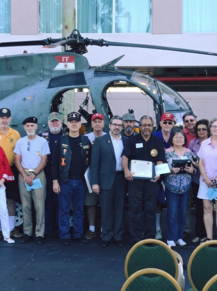 Grupo de personas frente a un helic&oacute;ptero, algunas con certificados, posando en un evento al aire libre.