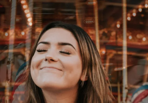 Mujer sonriendo con los ojos cerrados frente a un carrusel iluminado en un parque de diversiones.